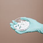 A gloved hand holds white pills in a petri dish, representing medical research.