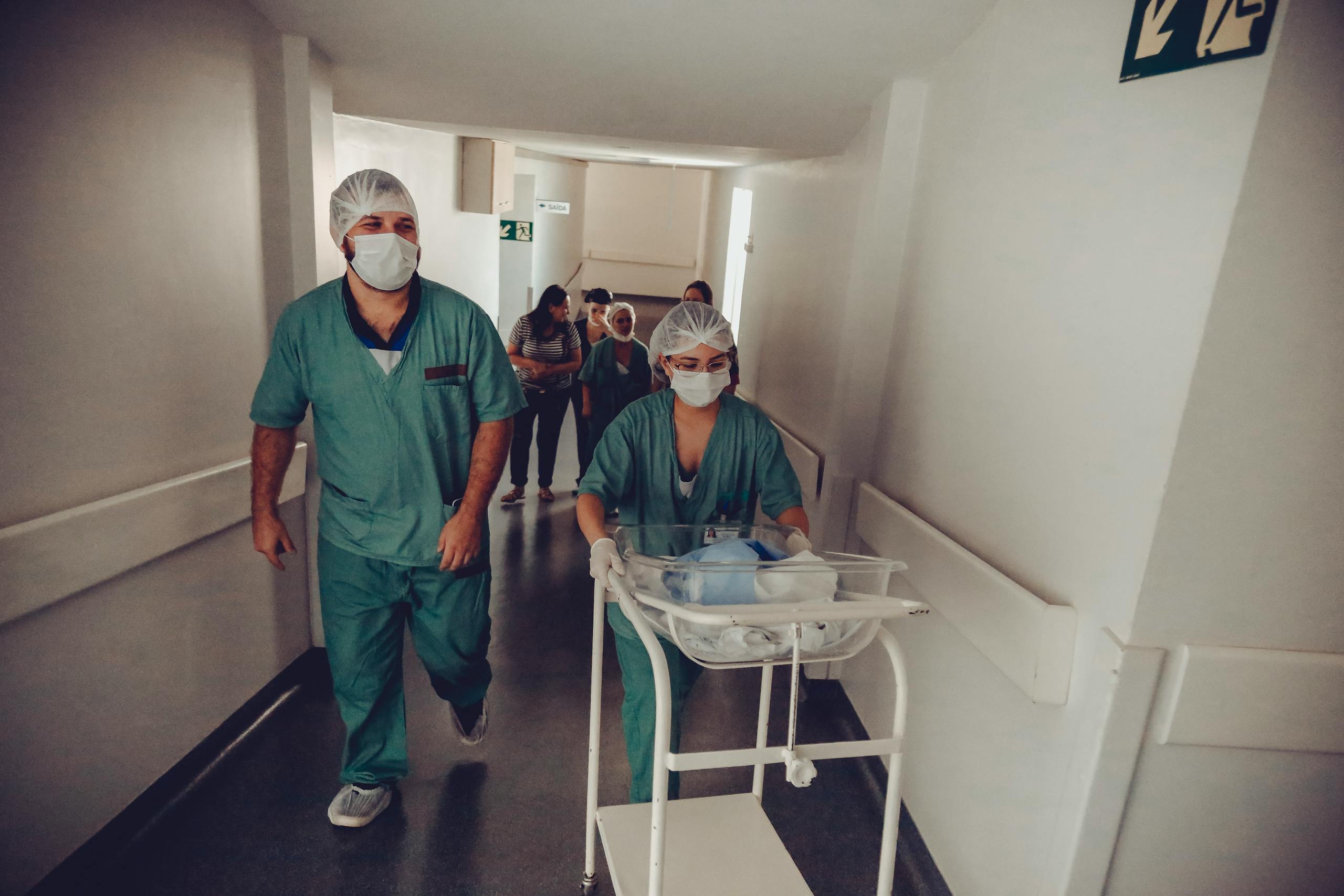 Healthcare workers in surgical attire transporting a newborn in a hospital corridor.